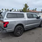 Silver Ford F-150 Lightning with a Ranch Fusion cap and roof rack, parked near a beige wall with palms.