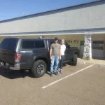 Black Toyota Tacoma TRD 4x4 with a Ranch Fusion cap, parked outside Canyon State RV with two people standing.