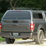 Gray Ford F-150 with a Ranch Fusion camper shell, parked on a gravel road with trees in the background.