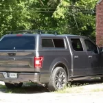 Gray Ford F-150 with a Ranch Fusion camper shell, parked near a brick building with trees in the background.