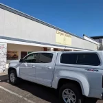 White Chevrolet Colorado with a Ranch Fusion camper shell from Canyon State RV, parked outside the store.